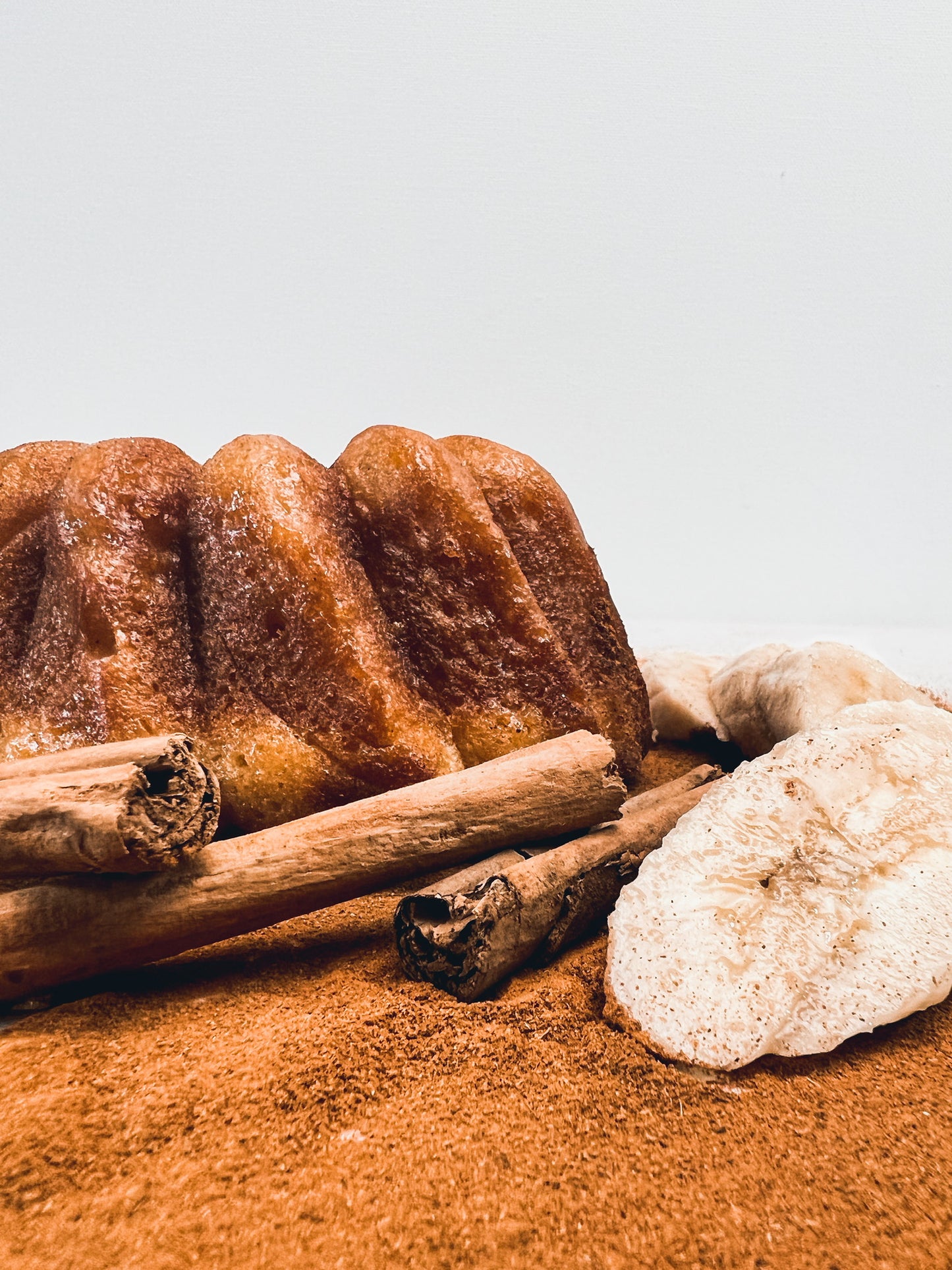 A cinnamon banana whiskey cake dusted with cinnamon powder, accompanied by fresh ripe banana slices and cinnamon sticks, presented on a white surface.