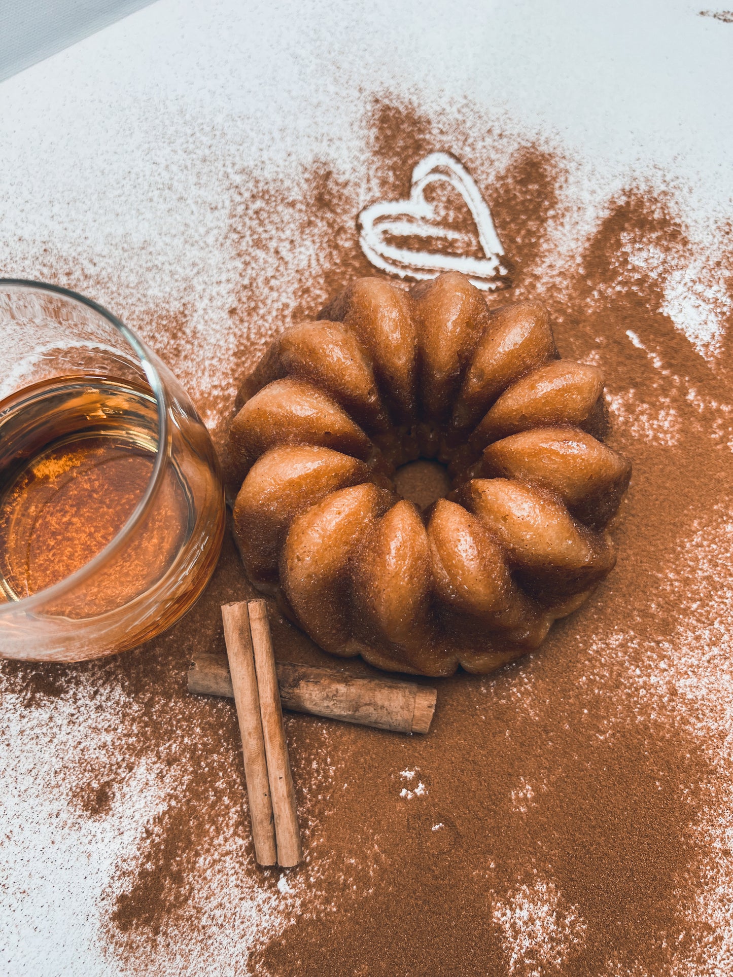 A cinnamon banana whiskey cake dusted with cinnamon powder, accompanied by a small glass jar of whiskey and cinnamon sticks, presented on a white surface.