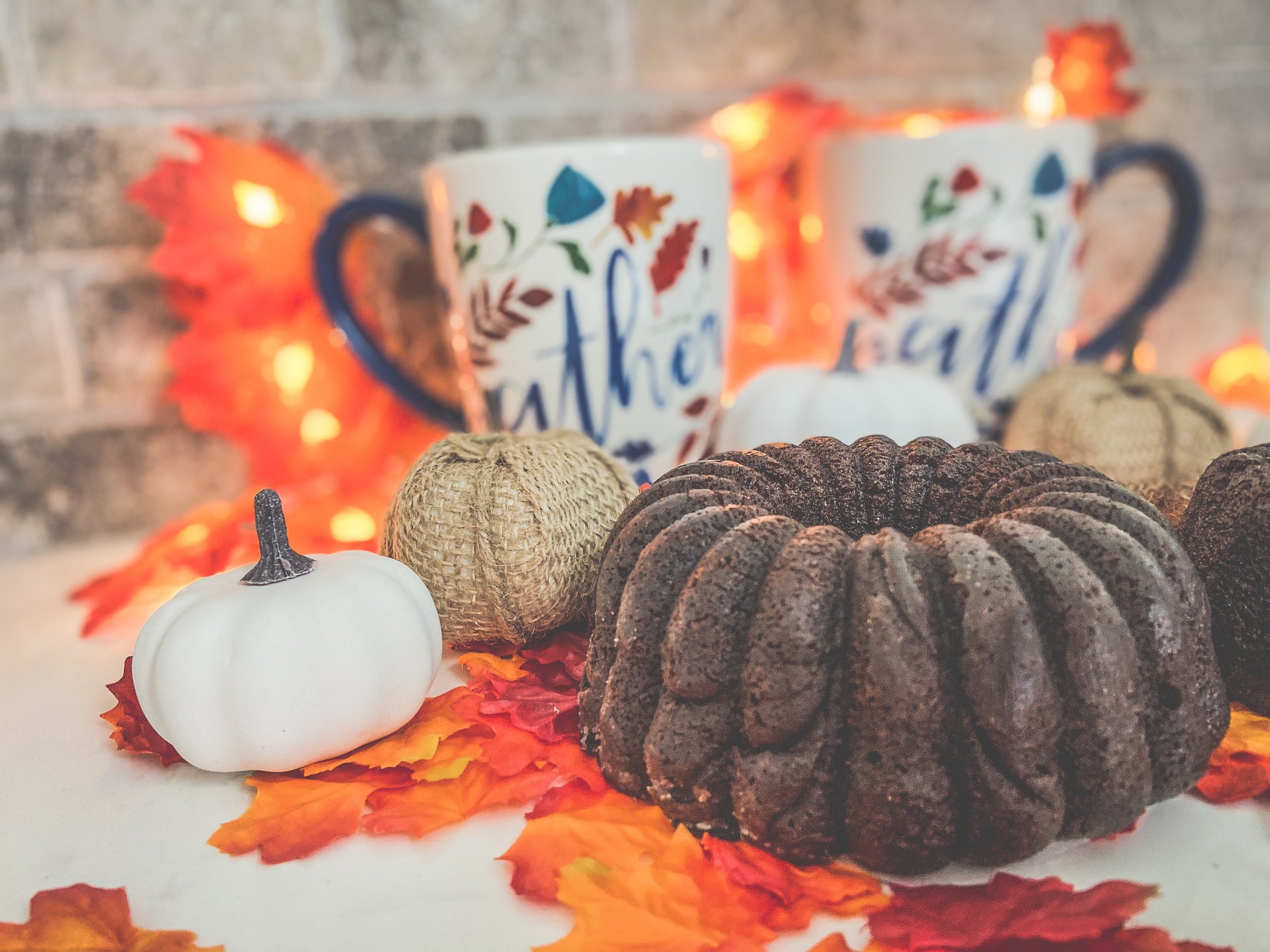 A photo of Peanut Butter Chocolate Fudge Whiskey Cake surrounded by Halloween and fall decor, including fake maple leaves, burlap wrapped pumpkins and fall aesthetic coffee mugs, on a white table with brick background.