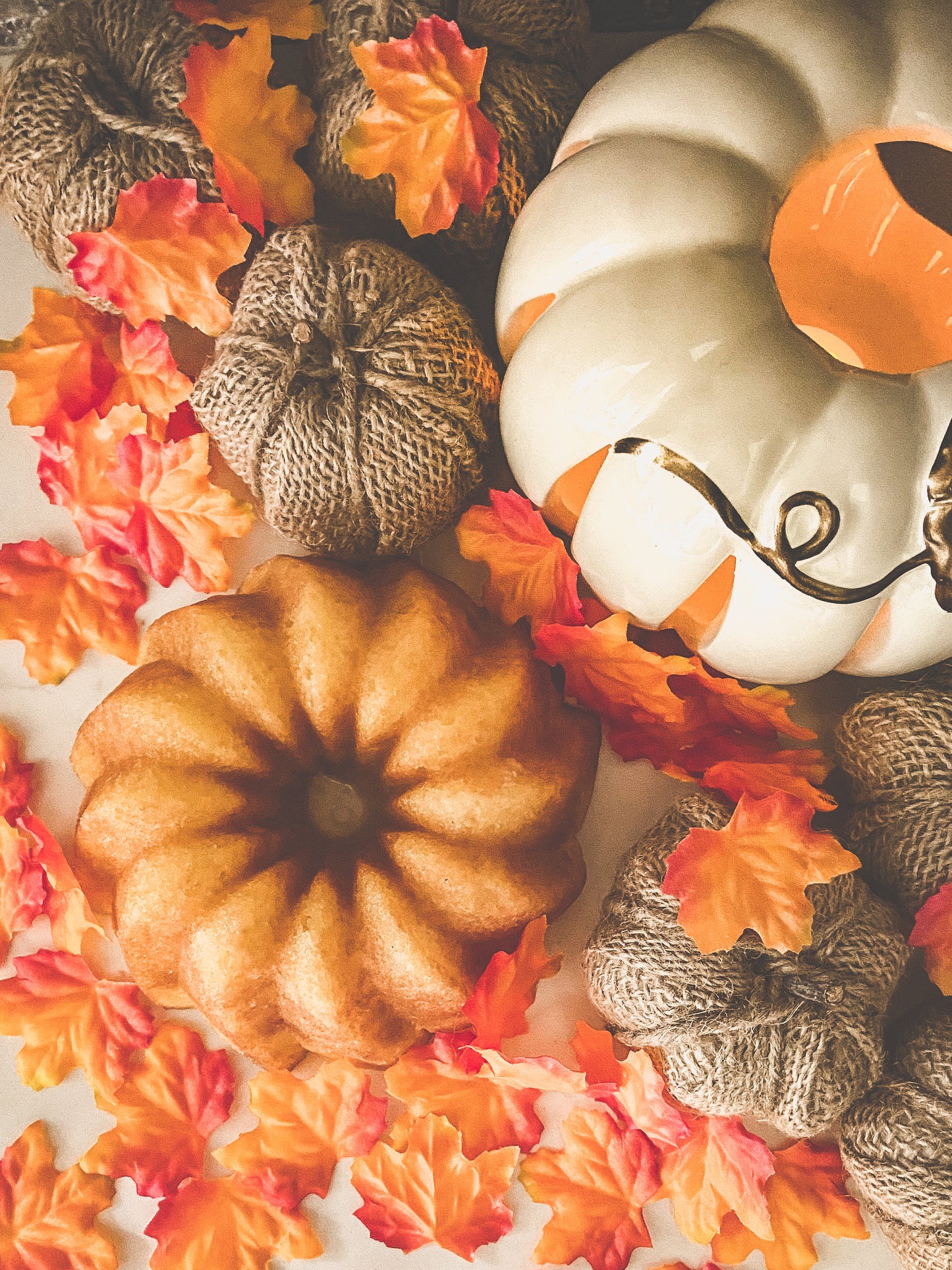 Photo of a vanilla rum cake accompanied by fall and thanksgiving decor, including fake maple leaves, burlap pumpkins and a white ceramic candle on  white background.