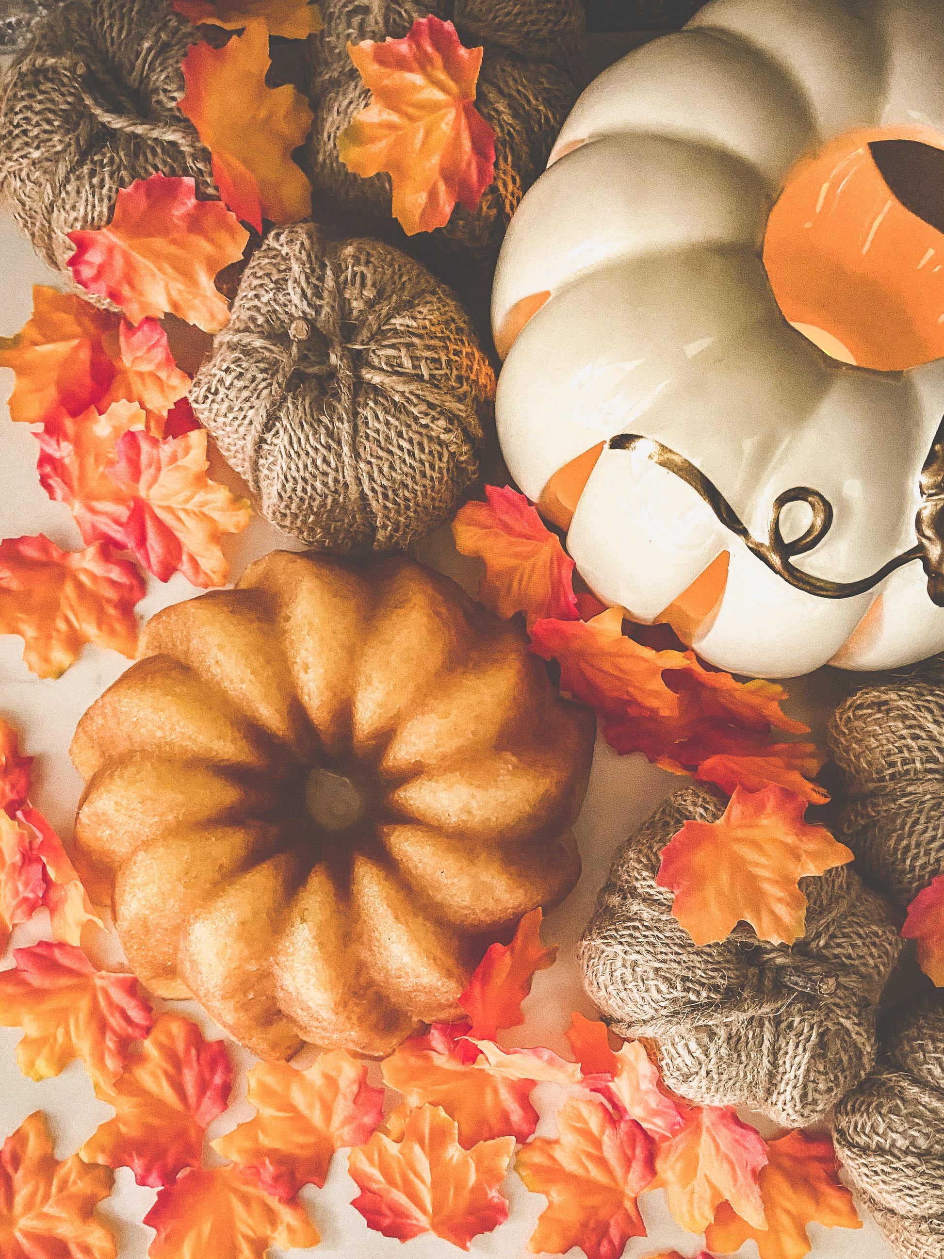 Photo of a vanilla rum cake accompanied by fall and thanksgiving decor, including fake maple leaves, burlap pumpkins and a white ceramic candle on  white background.